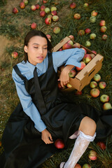 Portrait of a beautiful Asian woman in stylish clothes with apples in the garden. The girl is sitting on the ground and there are a lot of red apples around