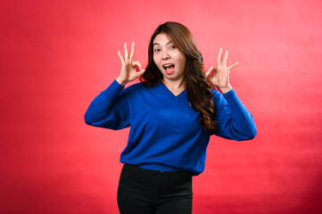 An Asian woman in a blue sweater and black pants poses in front of a red background, making double 