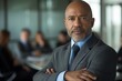 © N Joy Art  - Mature African American businessman with gray beard standing with arms crossed in a formal office setting. Colleagues discussing business matters in the background, showing confidence and authority.