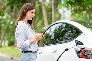 Young woman recharge EV electric vehicle battery from EV charging station and using smartphone online banking to pay for electricity in city park. Eco friendly vehicle travel with EV car. Exalt