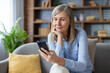 © Liubomir - Senior woman with gray hair using smartphone, sitting on sofa, looking thoughtful. She appears relaxed and engaged with technology, possibly checking messages or social media on phone.