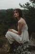 © StockUp - A thoughtful woman in a flowing white dress sits on a rock amidst lush greenery, creating a serene ambiance.