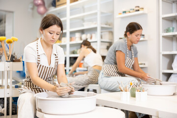 During master class, two young women and teen girl works with clay blank, processes with potter wheel, forms plate, vase product. Mastering ancient technique, craft as hobby