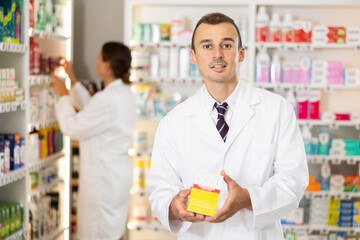 Wall Mural - Kind young apothecary holding a box with remedy standing against shelves full of medicine in drugstore