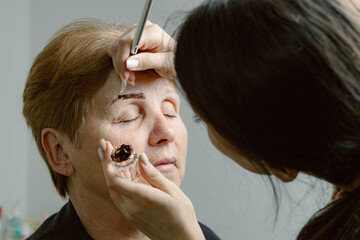 A cosmetologist paints the eyebrows of an elderly woman.