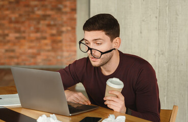 Poster - Sleepy Overworked Man At Laptop Holding Coffee Cup Trying To Work Sitting In Modern Office. Selective Focus
