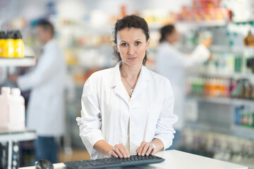 Wall Mural - Smiling middle-aged woman apothecary standing at the stand with her hand on keyboard in big pharmacy