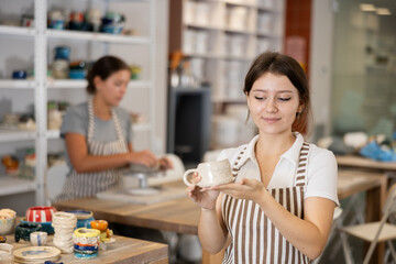 Wall Mural - Positive girl stands in pottery workshop and shows created work, finished decorative vase - jug. Female employee of workshop made individual product, completed task.