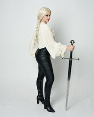 Wall Mural - Full length portrait of beautiful female model wearing vintage gothic fantasy blouse, leather pants, boots and blonde plaited hair, larp costume.  standing pose housing sword weapon in fighting action