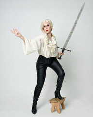 Poster - Full length portrait of beautiful female model wearing vintage gothic fantasy blouse, leather pants, boots and blonde plaited hair, larp costume.  standing pose housing sword weapon in fighting action