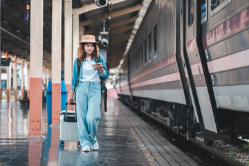 A young woman in casual attire walks along a train platform, checking her smartphone while pulling a suitcase.