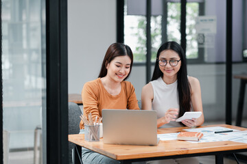 Two women working together at a desk with a laptop and documents in a modern office environment.