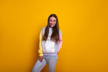 Smiling young woman poses against a vibrant yellow background, wearing pastel-colored hoodie and light jeans. Her playful and relaxed stance reflects a fun, casual, and modern style in this portrait