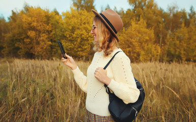 Wall Mural - Stylish happy smiling young woman with phone in autumn park, girl holding smartphone outdoors