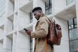 © SERHII - Stylish man with brown backpack checking phone in urban setting. Modern urban lifestyle and connectivity.