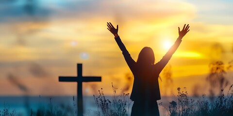 A woman is standing in a field with her hands raised in the air, and a cross is in the background. Concept of peace and hope, as the woman is praying or celebrating