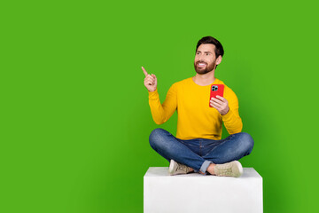 Poster - Full body photo of attractive man sit cube point look empty space hold device dressed yellow clothes isolated on green color background