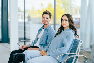 Young Caucasian man and woman sitting in modern waiting area with large windows. Couple travelers casually dressed in relaxed, comfortable clothing. Travel, leisure, and lifestyle concept.