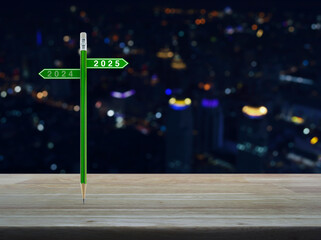 Wall Mural - 2025 and 2024 direction sign plate with green pencil on wooden table over blur colorful night light of modern office city tower and skyscraper, Business happy new year 2025 planning concept