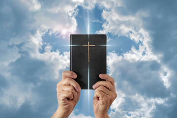 Wall Mural - Hands holding Holy Bible against the sky. Cross on a dark cover book. Sacred scripture.