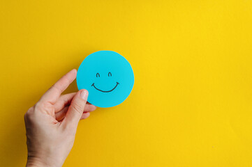 Happy face icon on a yellow background. A hand holds a blue smiley face icon on a bright yellow background. This cheerful image conveys positivity and optimism.