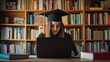 © Kamol. - Focused student wearing a graduation cap, studying online at home using a laptop and textbooks at a desk