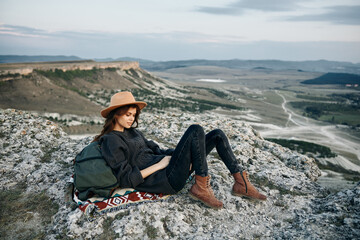 Wall Mural - adventurous woman sitting on mountain ledge with backpack and hat, enjoying breathtaking view