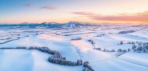 Wall Mural - Aerial view of a countryside blanketed in fresh snow, with distant mountains glowing under the warm colors of a winter sunset and a light snowfall enhancing the peaceful scene.