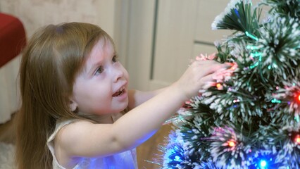 Child, little girl examines garland on Christmas tree. Child plays by Christmas tree in children room. Beautiful artificial christmas tree . happy childhood concept. Family play for Christmas holidays