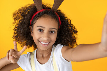 Happy young girl making selfie. Merry Christmas and New Year concept. African small kid daughter vlogging blogging online isolated over yellow background