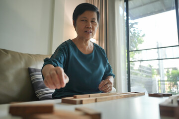 Wall Mural - Asian Senior woman concentrating while arranging wooden blocks on a white table in a living room.