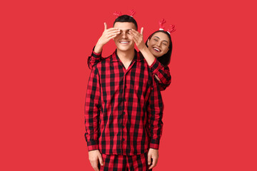 Canvas Print - Happy young woman in pajamas covering eyes of her husband with hands on red background. Christmas celebration