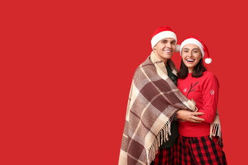 Poster - Happy young couple in Christmas pajamas and Santa hats wrapped in plaid on red background