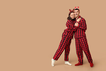 Wall Mural - Happy young couple in Christmas pajamas and with deer antlers hugging against beige background