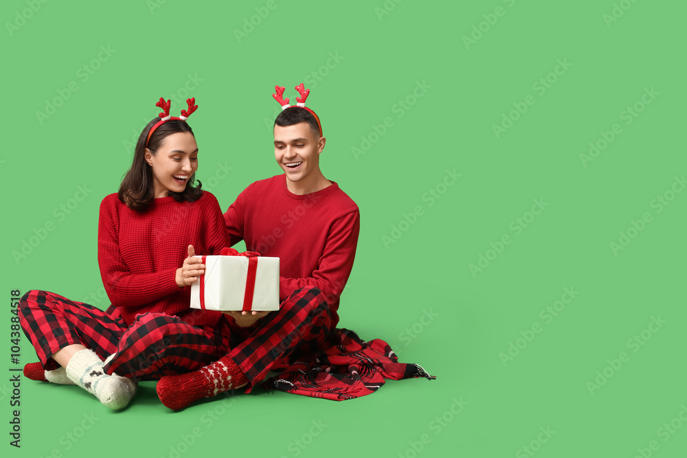 Happy young couple in Christmas pajamas and with gift on green background