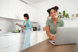 © Pixel-Shot - Young African-American lesbian woman using laptop and her girlfriend cooking in kitchen