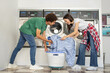 © Prostock-studio - A young couple is enjoying their time at a laundromat, happily loading a washing machine with clothes while sharing a moment of teamwork and laughter in a vibrant, clean environment.