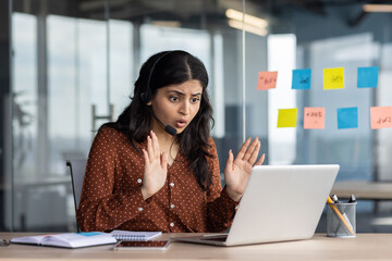 Wall Mural - Latina businesswoman in brown polka dot blouse looking surprised at laptop during video call in office. Professional interaction, remote work concept. Engaged communication through technology.