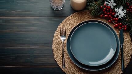 A beautifully arranged holiday table features two blue plates, silver cutlery, and decorative elements like candles and greenery, perfect for a winter gathering.