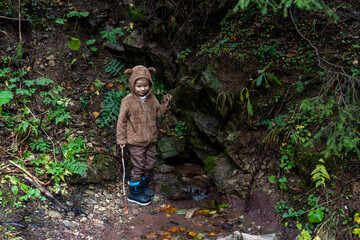 Young boy in a cozy bear onesie exploring a mountain forest near a small stream on a misty autumn day. Concept of childhood adventure, outdoor fun, and nature discovery. High quality photo