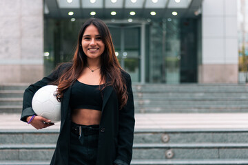 Beautiful brunette businesswoman posing, smiling with joy. Holding a soccer ball. Leaving work to train and play soccer. Outdoors. Sports and wellness advertising.