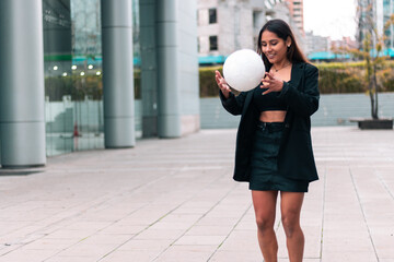 Beautiful brunette businesswoman holding and surprised with soccer ball. Wearing an office suit and skirt. Leaving work to train and play football. Outdoors. Sports and wellness advertising.