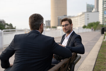 Business meeting of friends outdoors. Two men in suits are sitting on a bench near a city building with a laptop and talking. Partnership and corporate relations. Smiling partners discussing