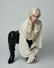 Sticker - full length portrait of beautiful female model, wearing vintage gothic fantasy ruffle blouse white shirt, leather pants, blonde hair holding knife weapon. sitting pose, isolated on studio background.