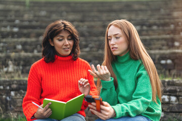 Two friends in brightly colored sweaters enjoy a study session outdoors on stone steps surrounded by nature