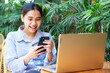 © M Alfan Setyawan - excited asian woman using smartphone with laptop on wooden table working from outdoor cafe with green plant