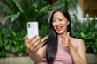 © bongkarn - A smiling Asian woman is enjoying a video call on her smartphone while sitting at an outdoor table.