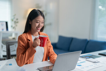 Young asian woman happily works from her home office, using her laptop to join an online meeting with her team, sipping coffee and smiling as she balances business tasks with relaxation