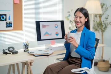 Young professional woman takes a coffee break while working from home, smiling and focused at her desk with a laptop and plant