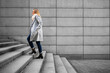 © Jacek Chabraszewski - Middle-aged blonde woman in gray coat, scarf, jeans and sunglasses walking up concrete stairs  against background of concrete slabs in city on autumn day. Businesswoman. Side view.